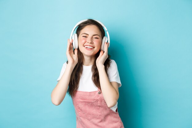 Linda chica caucásica en traje de primavera, escuchando música en auriculares, sonriendo complacido a la cámara, de pie sobre fondo azul.