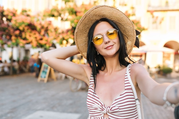Linda chica complacida con gafas de sol amarillas de moda pasar tiempo cerca del restaurante al aire libre esperando amigos y haciendo selfie