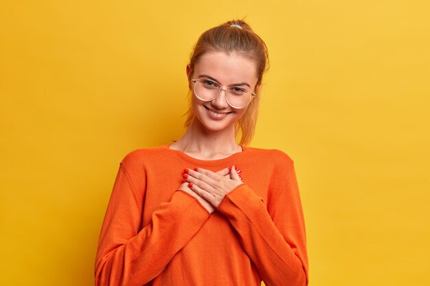 Linda chica complacida y satisfecha presiona las manos al corazón, aprecia un buen regalo, se ve agradecida por estar llena de amor y amabilidad sonríe tiernamente usa lentes ópticos, el jersey naranja se encuentra en el interior