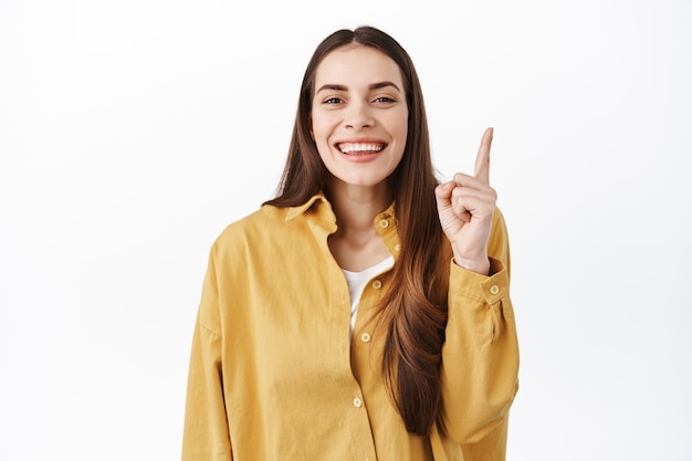 Linda chica coqueta se ríe, sonriendo y apuntando con el dedo hacia arriba, mostrando el anuncio arriba, la mejor oferta promocional, recomendando echar un vistazo, de pie sobre una pared blanca