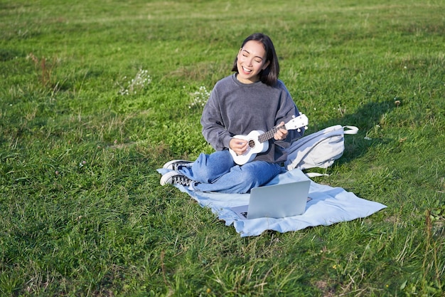 Linda chica coreana músico se sienta en el parque toca el ukelele y canta busca acordes y tutoriales en lapt