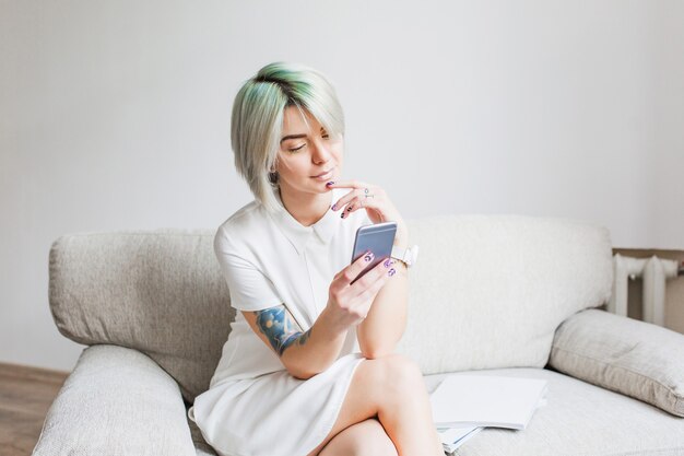 Linda chica con corte de pelo corto blanco con vestido blanco está sentada en el sofá en el estudio. Ella está escribiendo en el teléfono y sonriendo.