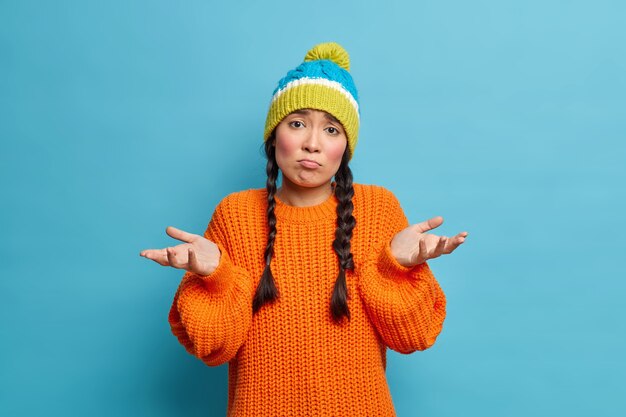 Linda chica despistada se encoge de hombros y se ve dudosa al frente extiende las palmas tiene expresión de cara dudosa usa gorro de punto y suéter no sabe o no puede ayudar aislado sobre una pared azul