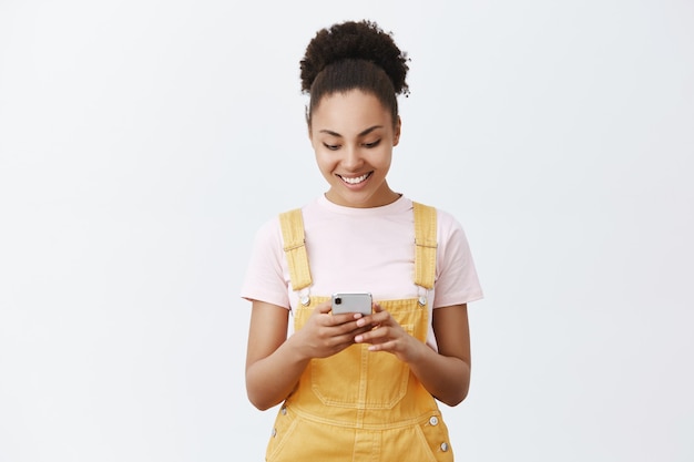 Linda chica despreocupada haciendo horario para mañana con nueva aplicación. Retrato de encantadora mujer urbana con piel oscura con un mono amarillo, sosteniendo el teléfono inteligente, sonriendo a la pantalla mientras escribe el mensaje