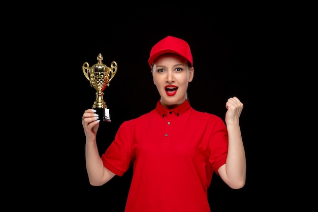 Linda chica del día de la camisa roja emocionada por un precio dorado en una gorra roja con camisa y lápiz labial brillante