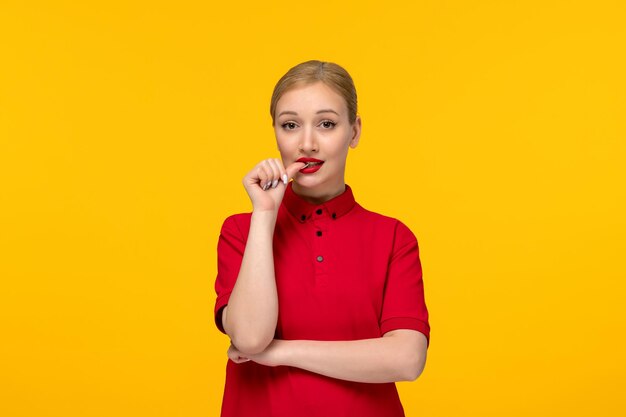 Linda chica del día de la camisa roja mordiéndose las uñas con una camisa roja y lápiz labial sobre un fondo amarillo