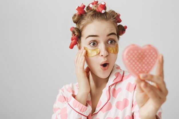 Linda chica emocionada en rizadores de pelo y parches en los ojos mirando en el espejo