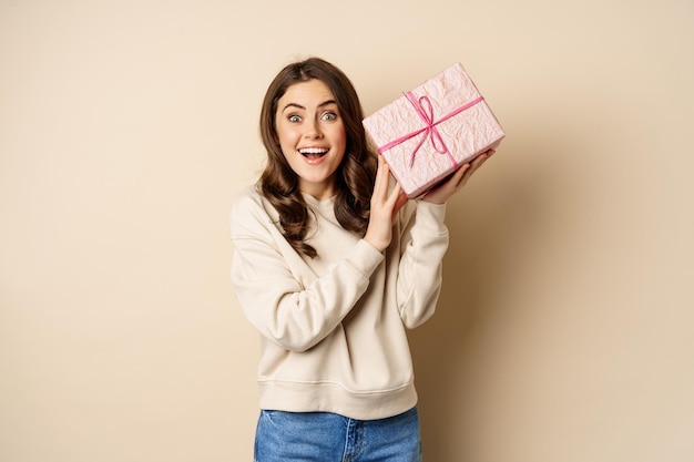 Linda chica emocionada sacudiendo la caja con el regalo adivinando qué hay dentro presente de pie feliz contra el fondo beige...