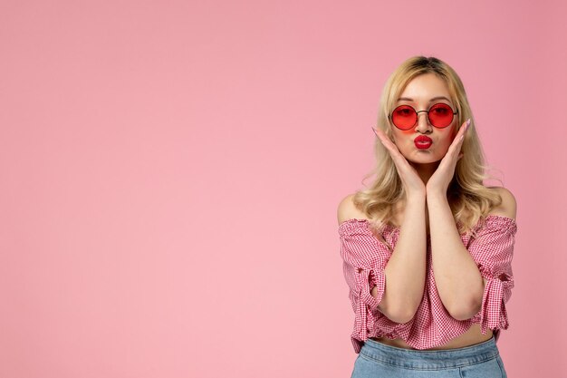 Linda chica encantadora jovencita con gafas rojas en blusa rosa enviando besos y sosteniendo la cara