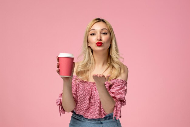 Linda chica encantadora jovencita con lápiz labial rojo en blusa rosa enviando besos y vaso de papel rosa