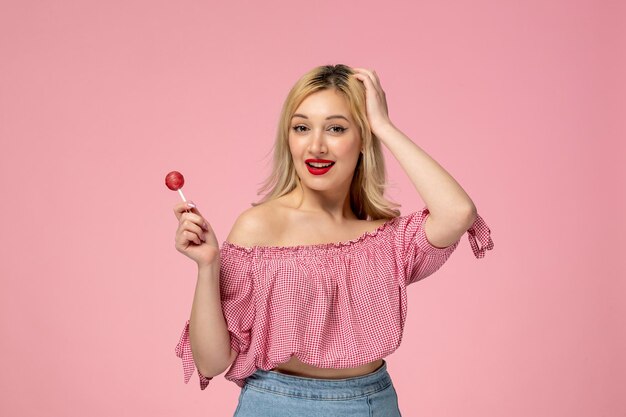 Linda chica encantadora jovencita con lápiz labial rojo en blusa rosa sosteniendo la cabeza y una piruleta rosa