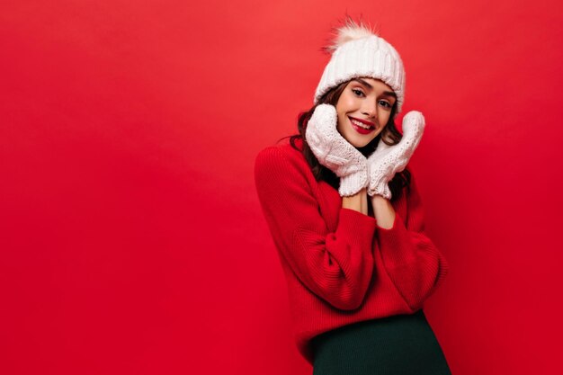 Linda chica encantadora en suéter brillante sombrero de punto y sonrisas de gatitos Mujer encantadora en traje cálido posa sobre fondo rojo