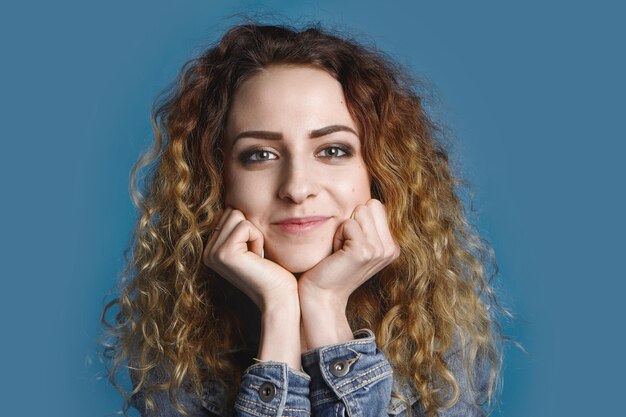 Linda chica encantadora de unos veinte años descansando la barbilla en sus manos con interés mientras escucha una historia genial. Sonriente mujer joven alegre en denim posando en la pared azul