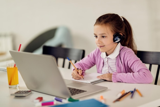 Linda chica escribiendo notas mientras aprende en línea sobre una computadora portátil en casa