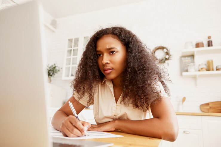 Foto gratuito linda chica estudiante afroamericana con mirada seria haciendo los deberes en la mesa de comedor, sentado frente a la computadora portátil abierta, haciendo notas con lápiz. elegante mujer negra con gadget electrónico para trabajo remoto