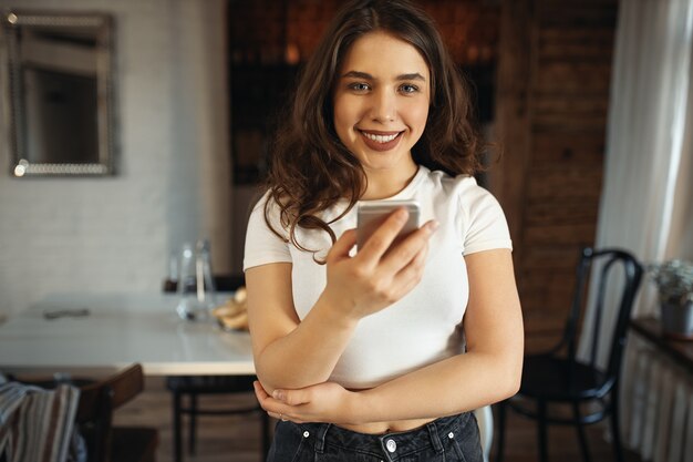 Linda chica estudiante en top blanco sosteniendo teléfono celular, enviando mensajes de texto a amigos a través de la aplicación messengaer, haciendo planes para el fin de semana