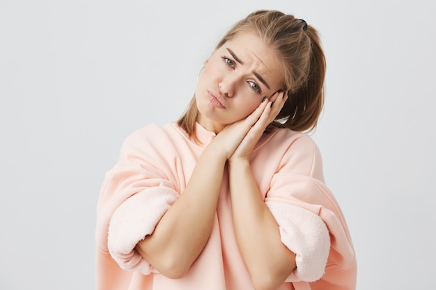 Linda chica europea elegante vistiendo sudadera rosa y mirando con expresión cansada, tomados de la mano debajo de la barbilla. Bastante cansado y somnoliento hembra descansando su cabeza sobre sus manos.