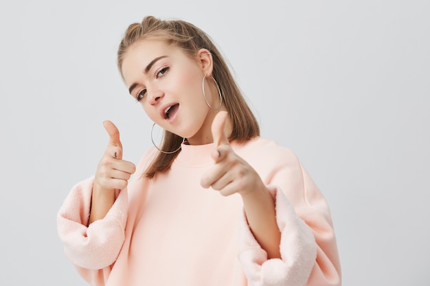 Linda chica europea juguetona y elegante con la boca abierta y el pelo liso y rubio con sudadera rosa de manga larga, posando, burlándose, apuntando con sus dedos índices