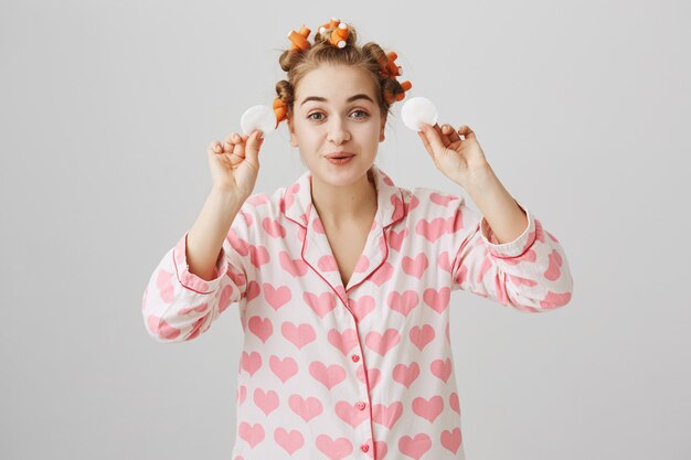 Linda chica feliz en ropa de dormir y rizadores de pelo limpie el maquillaje con almohadillas de algodón