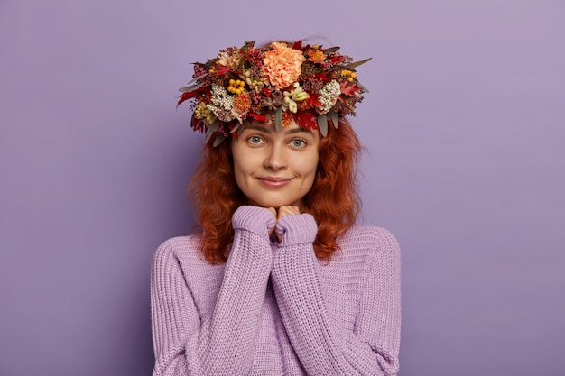 Linda chica femenina con cabello rojo ondulado, aprieta las manos juntas de manera suplicante, anticipa un gran momento, usa un suéter de punto de gran tamaño, corona otoñal