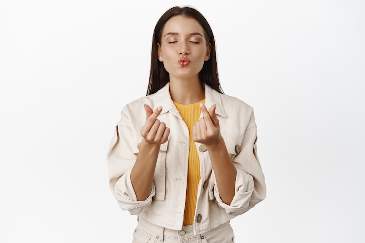 Foto gratuito linda chica femenina cierra los ojos, besa los labios y el gesto de los corazones de los dedos, te amo, como signo, pensando en algo romántico, de pie contra el fondo blanco