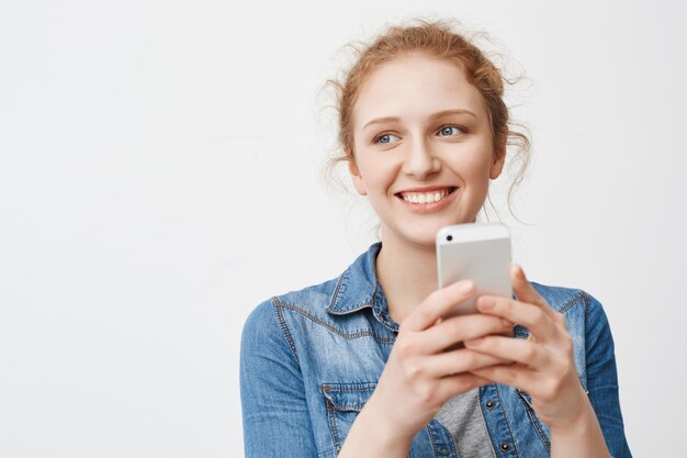 Linda chica femenina de jengibre con peinado desordenado mirando a un lado y sonriendo ampliamente mientras sostiene el teléfono inteligente