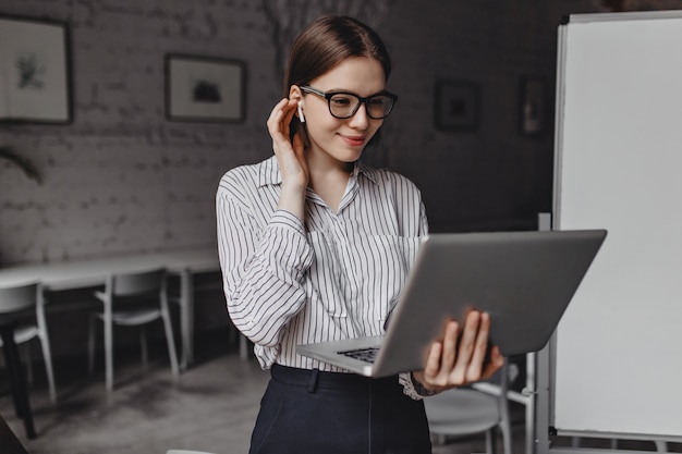 Linda chica con gafas elegantes sonríe, se pone el auricular inalámbrico y sostiene la computadora portátil abierta en el fondo del tablero de la oficina.