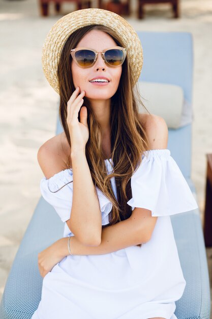 Linda chica con gafas de sol marrones y sombrero de paja sonríe ampliamente y sostiene su mano cerca de la barbilla