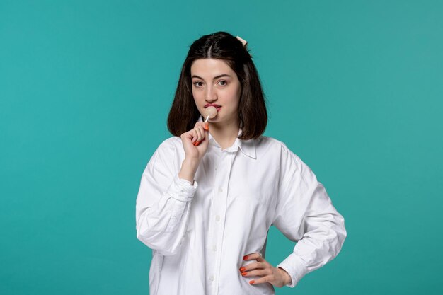 Linda chica guapa morena joven dulce con camisa blanca lamiendo una piruleta blanca