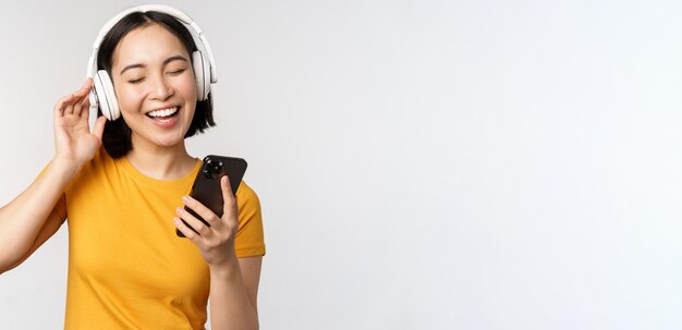 Linda chica japonesa en auriculares mirando el teléfono móvil y sonriendo usando la aplicación de música en el teléfono inteligente de pie contra el fondo blanco