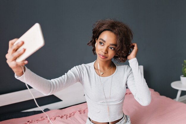 Linda chica joven, adolescente tomando selfie, escuchando música en la cama con alfombra rosa en la habitación con pared gris. Vistiendo camiseta gris claro con mangas largas, triángulo colgante.