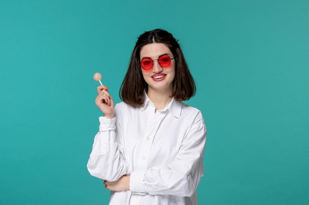 Linda chica joven bastante adorable con camisa blanca sosteniendo una piruleta blanca con gafas rojas