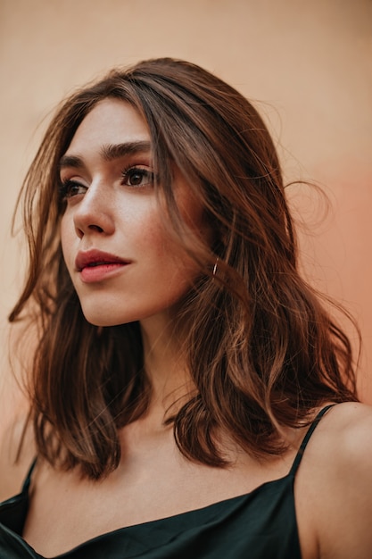 Linda chica joven con cabello ondulado morena, ojos oscuros y maquillaje elegante posando en vestido de correa contra la pared de la pared de melocotón y mirando a otro lado