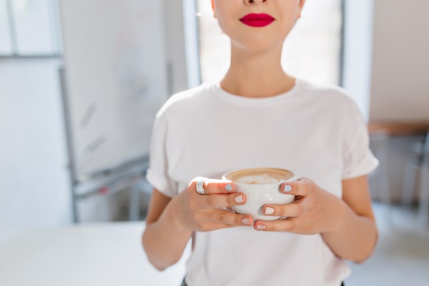 Linda chica con labios rojos y manicura de moda sosteniendo una taza de sabroso café disfrutando del sabor en un día ajetreado