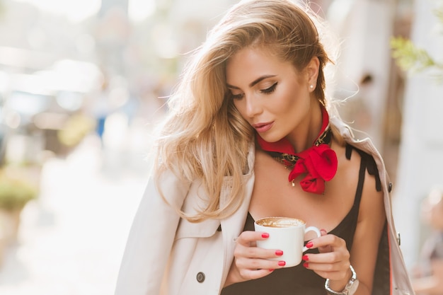 Linda chica con maquillaje de moda relajante en un día soleado y bebiendo café con leche con los ojos cerrados. Retrato al aire libre de hermosa mujer bronceada con cabello rubio posando en abrigo con taza de café.