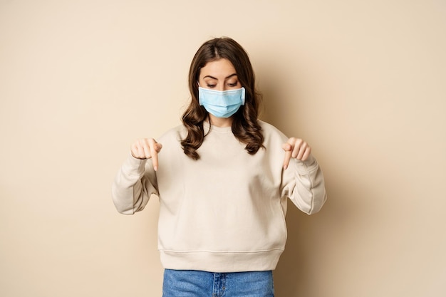 Linda chica con mascarilla médica, señalando con el dedo y mirando hacia abajo, mostrando un anuncio debajo, de pie contra un fondo beige