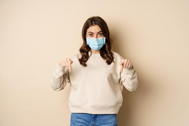 Linda chica con mascarilla médica, señalando con el dedo y mirando hacia abajo, mostrando un anuncio debajo, de pie contra un fondo beige