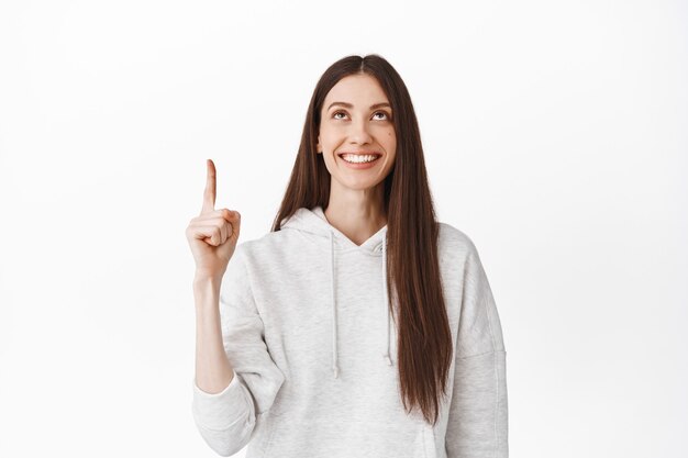 Linda chica milenaria mirando algo arriba, señalando con el dedo en la parte superior y sonriendo complacida, encontró un anuncio promocional genial, mostrando un enlace o logotipo, de pie contra una pared blanca