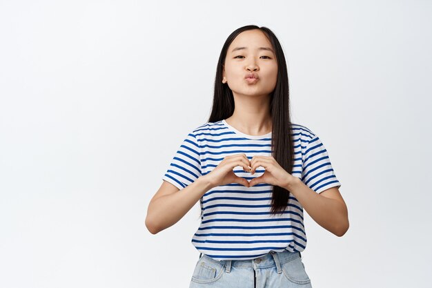 Linda chica morena asiática fruncir los labios, mostrando besos en la cara y el signo del corazón, haciendo una confesión de amor, de pie en ropa de verano en blanco