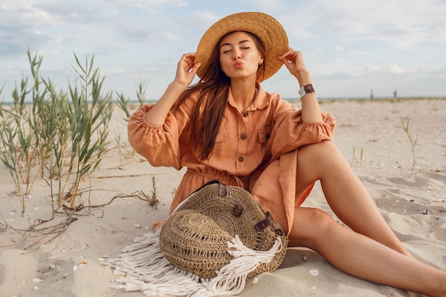 Linda chica morena enviar beso y relajarse en la playa. Vistiendo ropa de lino de moda de verano. Sombrero de paja y bolso boho.