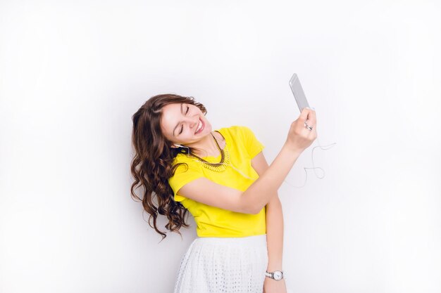 Linda chica morena escuchando música con auriculares en el teléfono inteligente se divierte y toma un selfie. Ella sonríe ampliamente. Tiene el pelo castaño largo y ondulado. Viste camiseta amarilla y falda blanca.