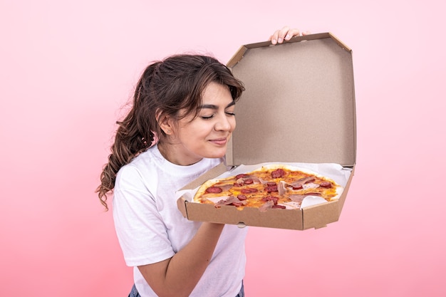 Linda chica morena oliendo pizza de una caja de entrega abierta, fondo rosa.