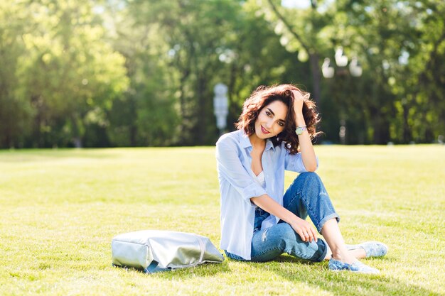 Linda chica morena con el pelo corto posando sobre la hierba en el parque. Viste camiseta blanca, camisa y jeans, zapatos. Ella está sonriendo a la cámara a la luz del sol.