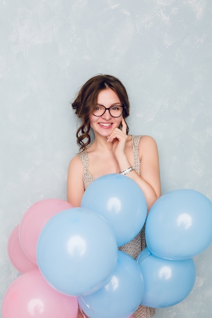 Linda chica morena de pie en un estudio, sonriendo ampliamente y sosteniendo globos azules y rosas. . Ella toca sus lentes