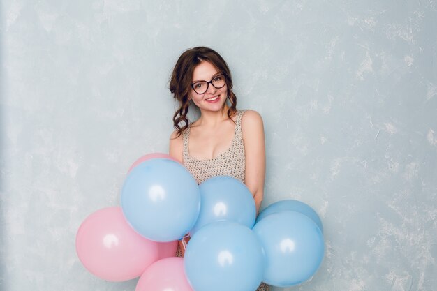 Linda chica morena de pie en un estudio, sonriendo ampliamente y sosteniendo globos azules y rosas.