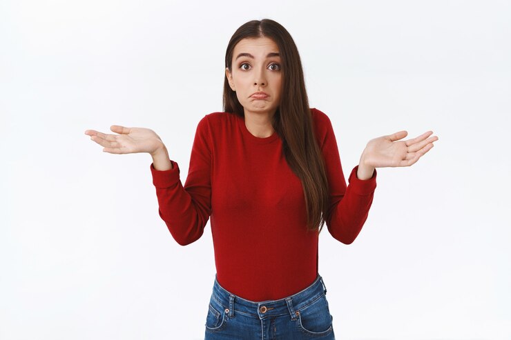 Foto gratuito una linda chica morena preocupada y desorientada con un suéter rojo no tiene ni idea de lo que está sucediendo, extiende las manos hacia los lados y frunce los labios, menea la cabeza sin darse cuenta, no puede decidir pararse indeciso