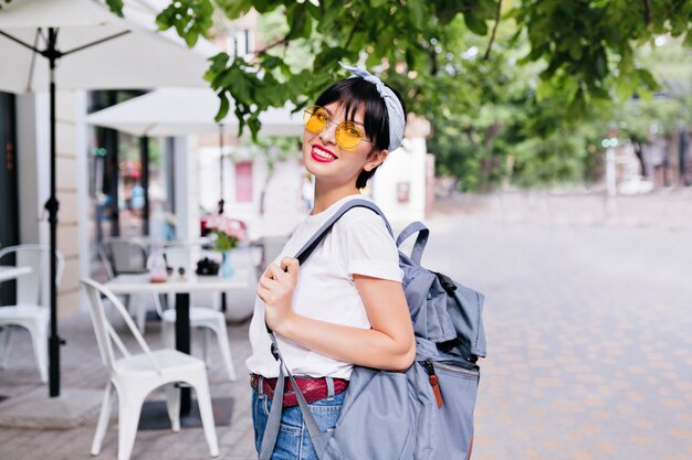 Linda chica morena sonriente con gafas de sol amarillas y cinturón de cuero con mochila mientras explora la ciudad