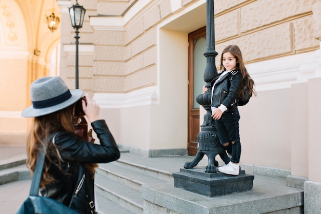 Linda chica morena con zapatillas blancas y pantalones de mezclilla sosteniendo por pilar, mientras que la madre toma una fotografía de pie frente a ella. Mujer joven elegante que lleva el bolso de cuero y la cámara que hace la foto.