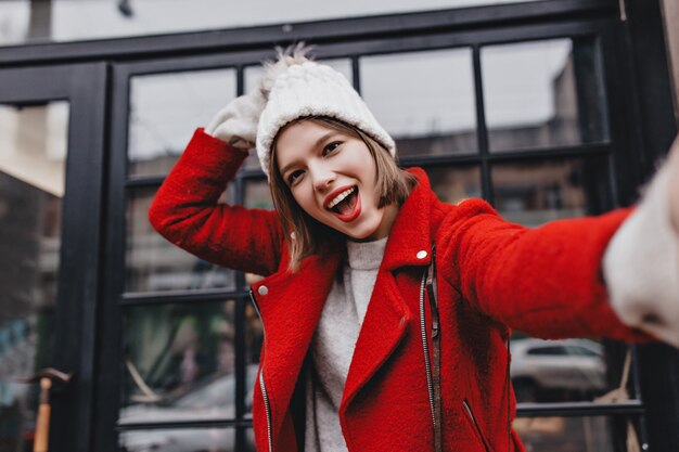 Linda chica de ojos marrones de buen humor hace selfie. Retrato de niña con chaqueta roja y gorro de punto blanco.