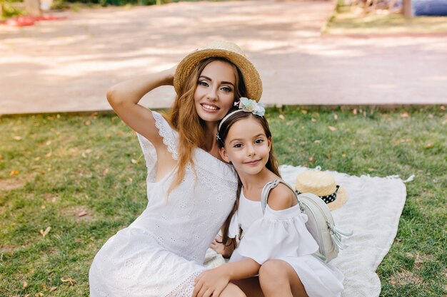 Linda chica de ojos oscuros con mochila de cuero posando sobre una manta con elegante joven madre lleva sombrero de paja. Retrato al aire libre de mujer refinada en vestido de encaje abrazando a hija con cinta en el pelo.